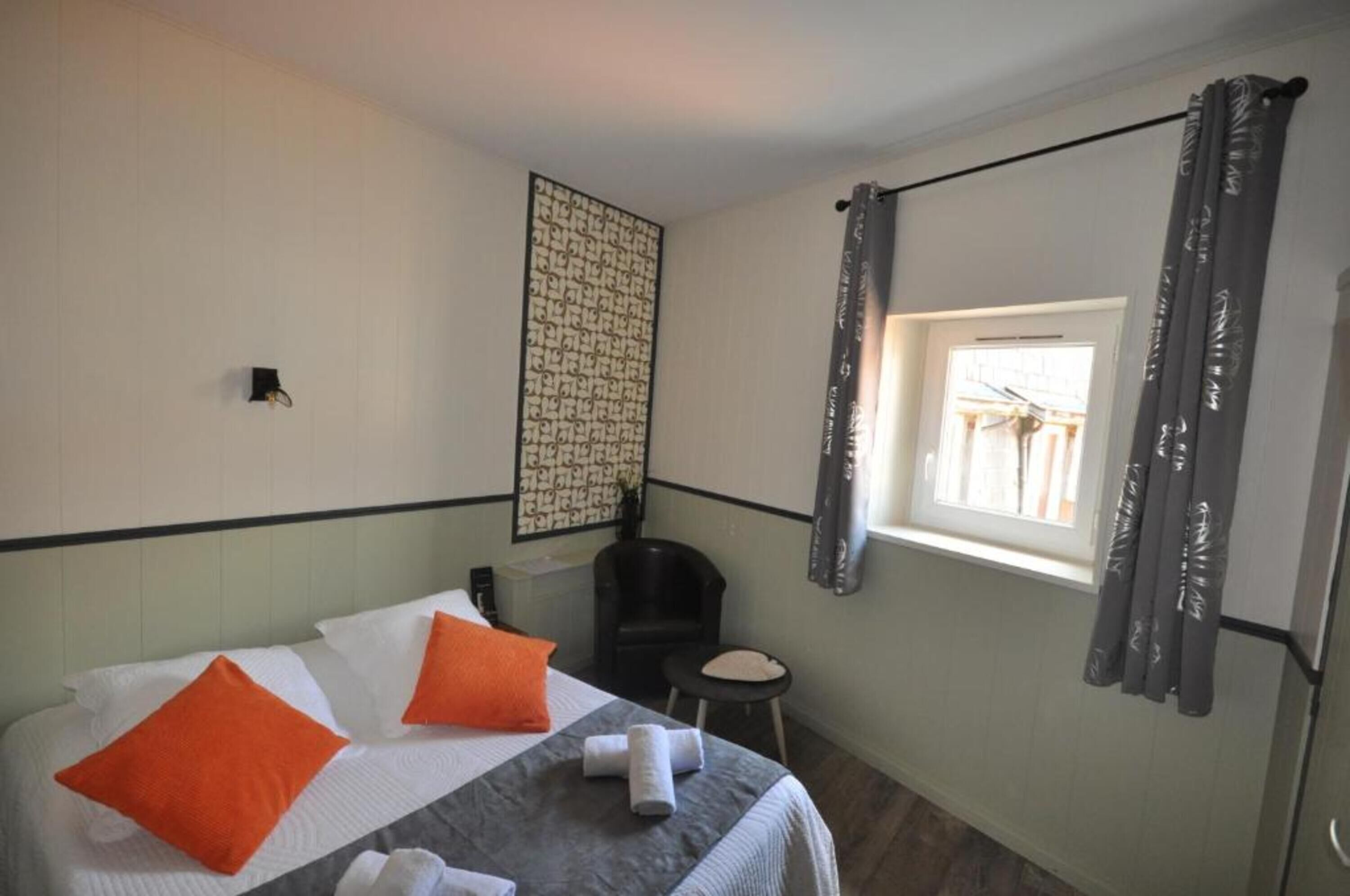 Hotellihuone, jossa on sänky, pieni jakkara, tuoli, ikkuna verhoilla ja seinään kiinnitetty valaisin.