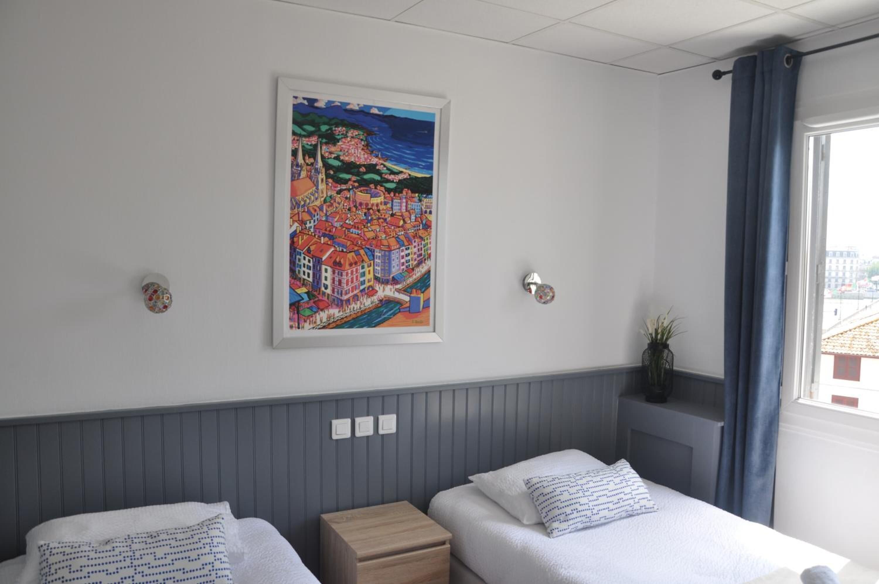 Een hotelkamer met twee bedden, een stadsgezicht op een schilderij, een raam met gordijnen en een plant op de vensterbank.