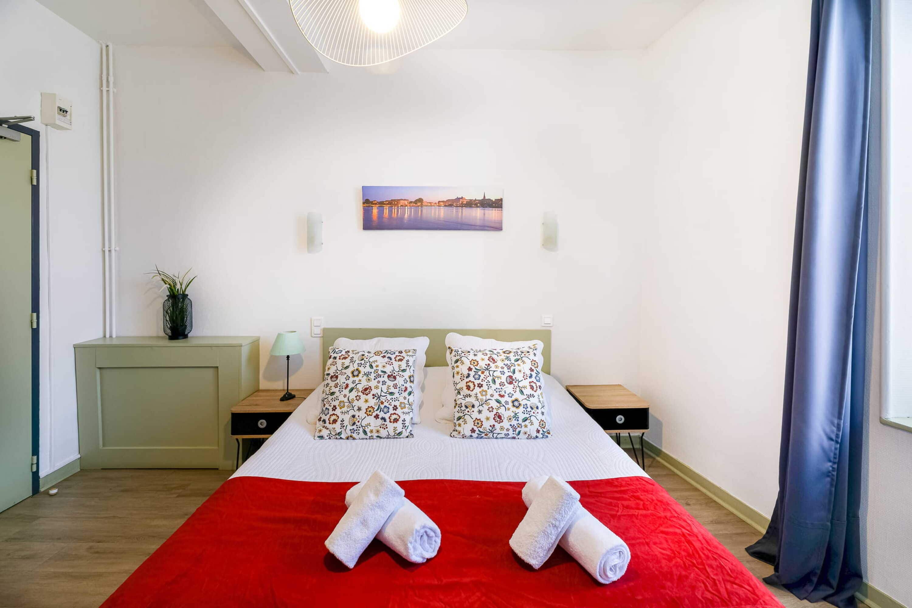 Habitación de hotel con cama, dos mesitas de noche, colcha roja, dos almohadas, un cuadro en la pared y una ventana con cortinas.