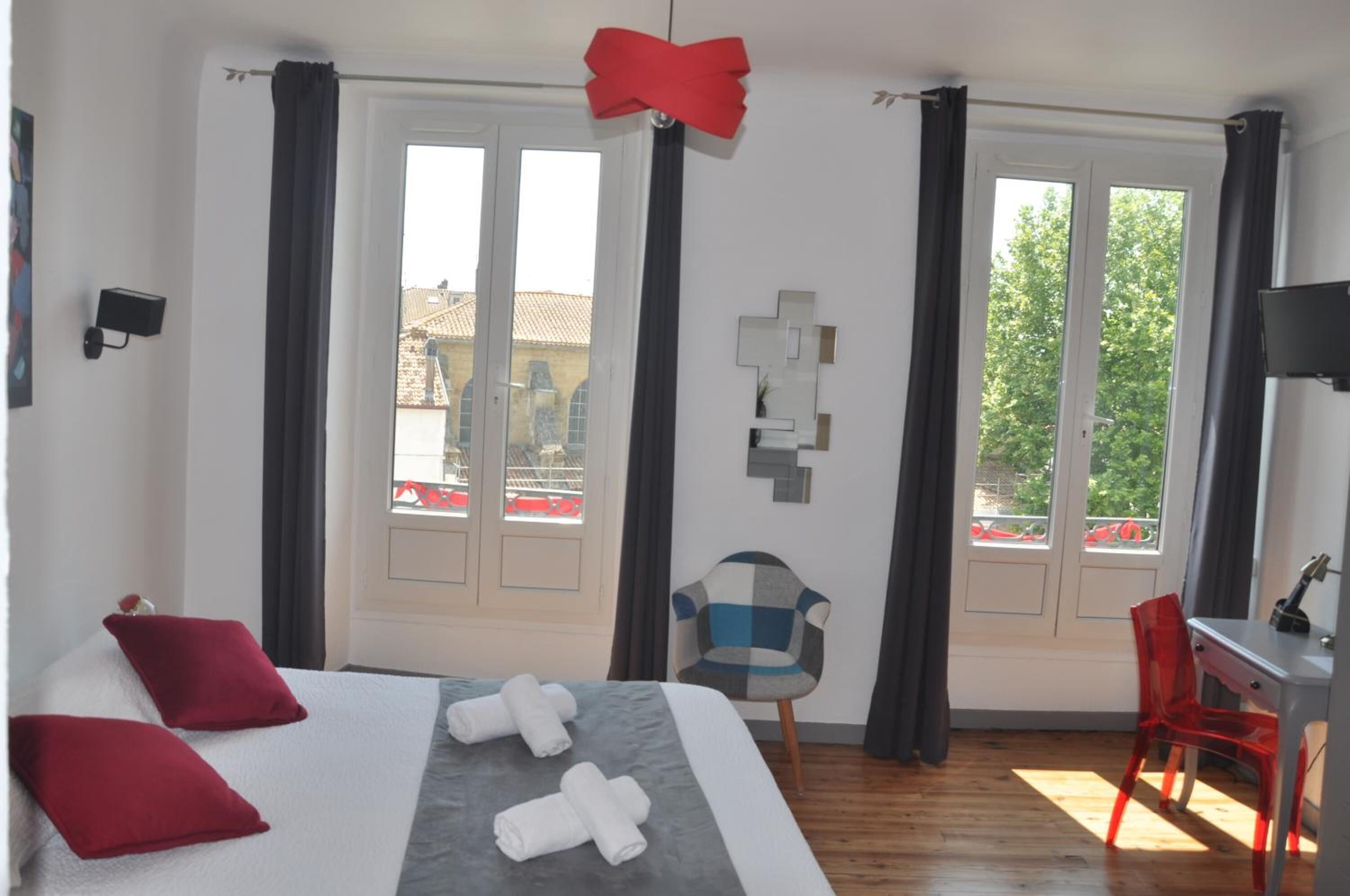 A hotel room with a bed, two red pillows, a desk with a chair, and large windows with curtains.
