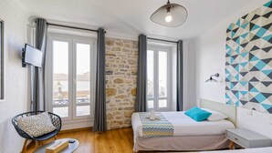 A hotel room with a stone wall, two beds with patterned bedding, a TV mounted on the wall, and a balcony view. - Hôtel Côte Basque (Bayonne)
