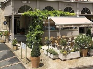 Un espacio al aire libre con mesa y sillas cubiertas, plantas en macetas e indicación de hotel y restaurante.