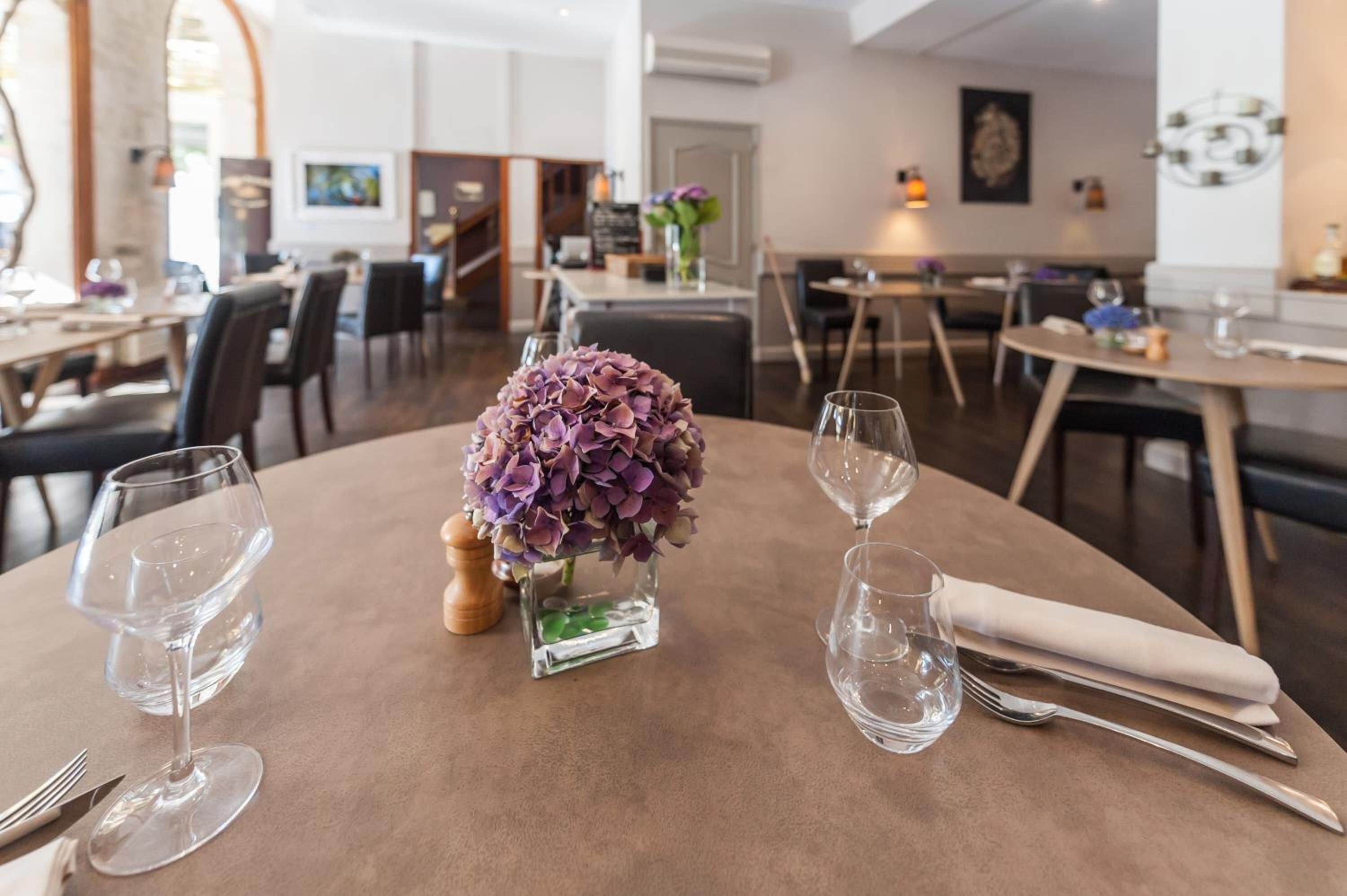 A dining area with round tables, each set with wine glasses, cutlery, and a centerpiece of purple flowers.