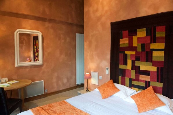A hotel room with a bed, a desk, a chair, a mirror, and a wall with a colorful quilt.