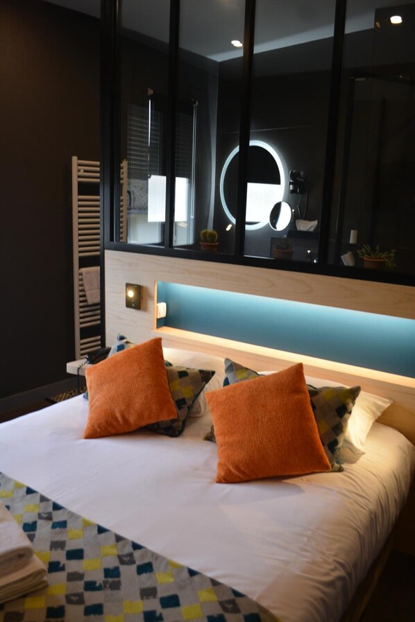 A modern hotel room with a large bed, two orange pillows, a blue headboard, and a large mirror with a circular light. - Hôtel les Glycines (Melle)