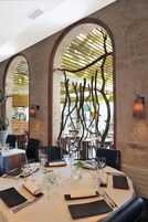 Un restaurant aux murs de pierre, aux fenêtres en arc de cercle et dont la décoration est centrée sur une branche d’arbre. Les tables sont dressées avec des verres à vin, des assiettes et des couverts.