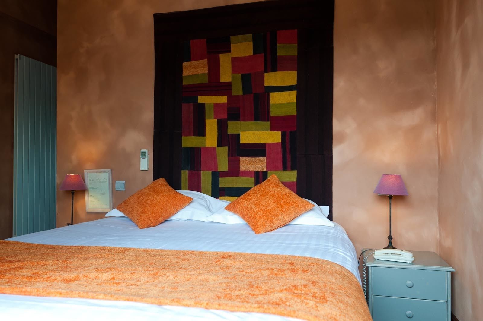Habitación de hotel con cama, almohadas naranjas, un tapiz con diseño y dos lámparas de noche.