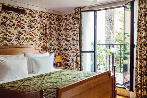 Un dormitorio con papel tapiz floral, una cama de madera, colcha verde, una mesita de noche con lámpara y un balcón con plantas.