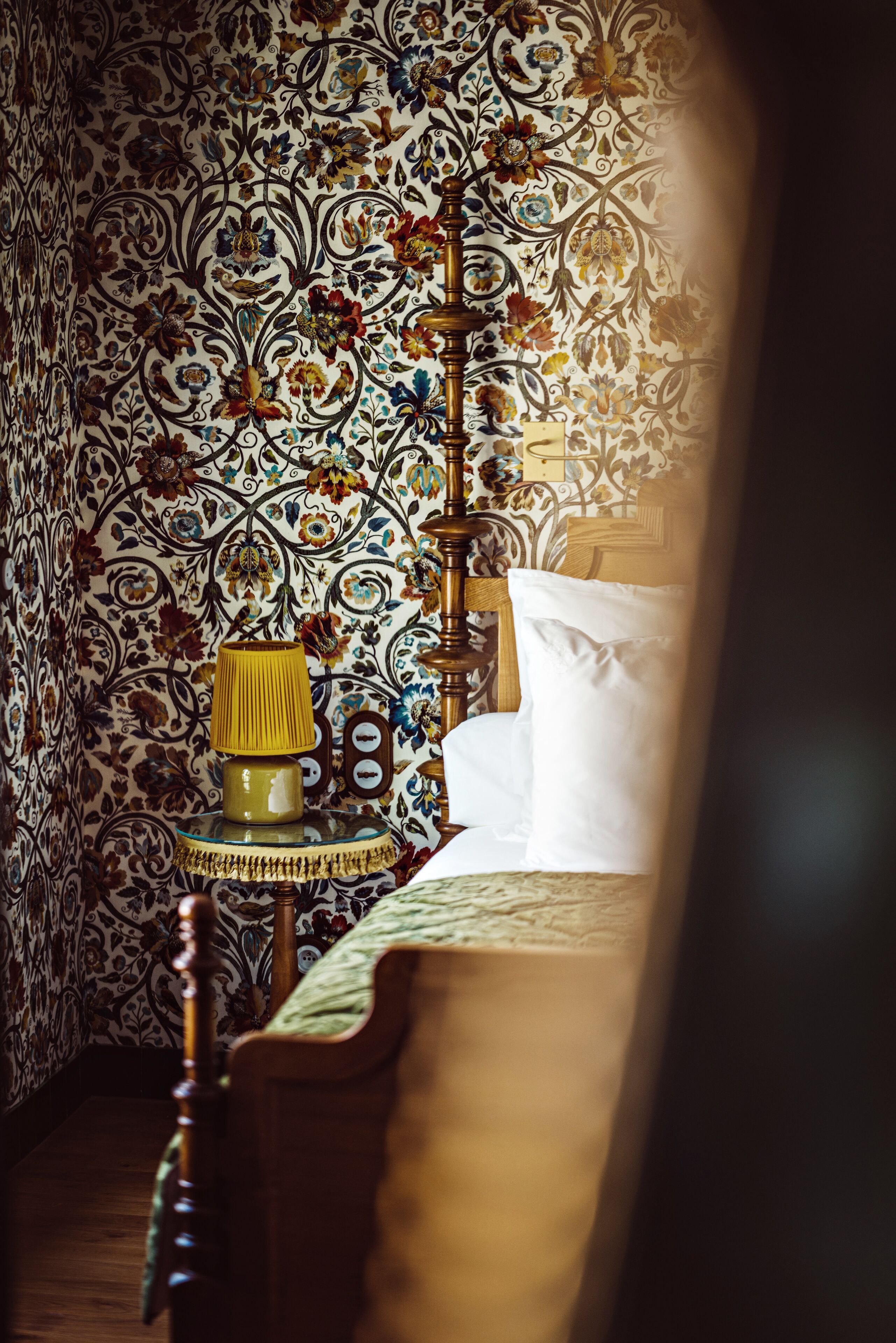 A bedroom with a bed, a bedside table with a lamp, and a wall with a floral pattern.