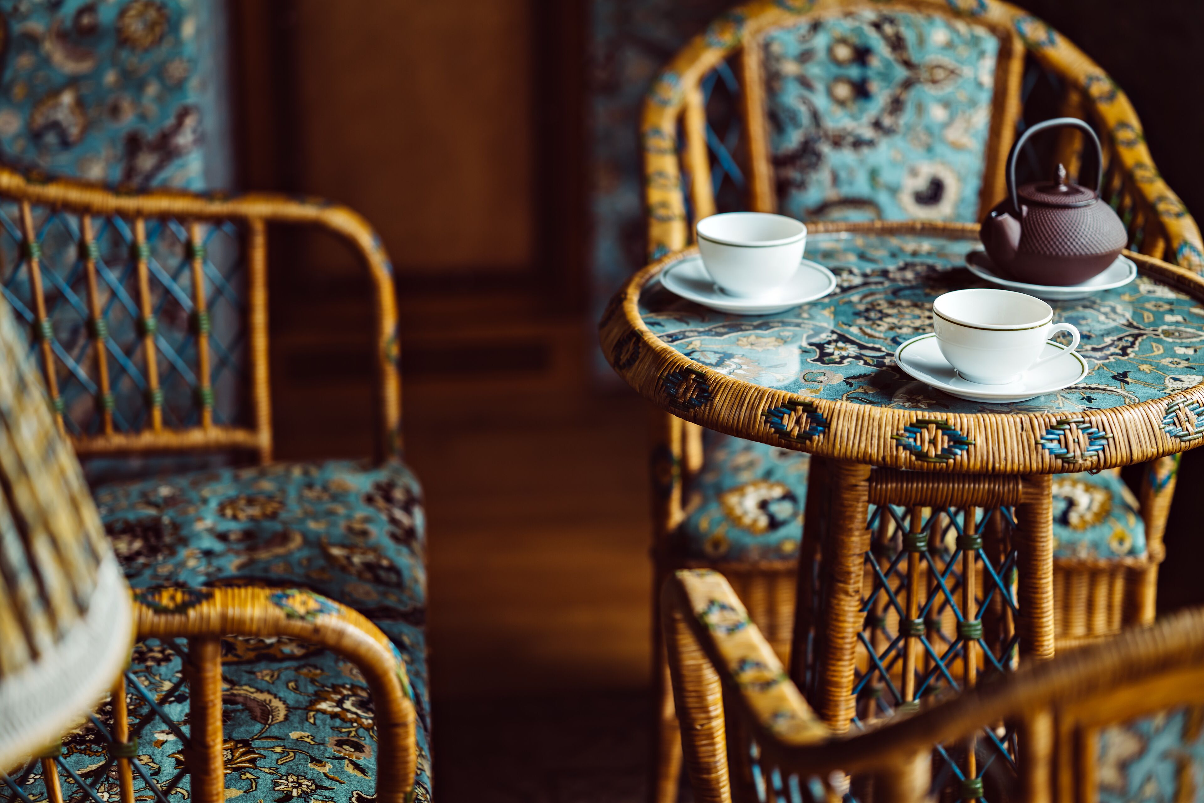 A wicker chair and table with a patterned tablecloth, a teapot, and two cups.