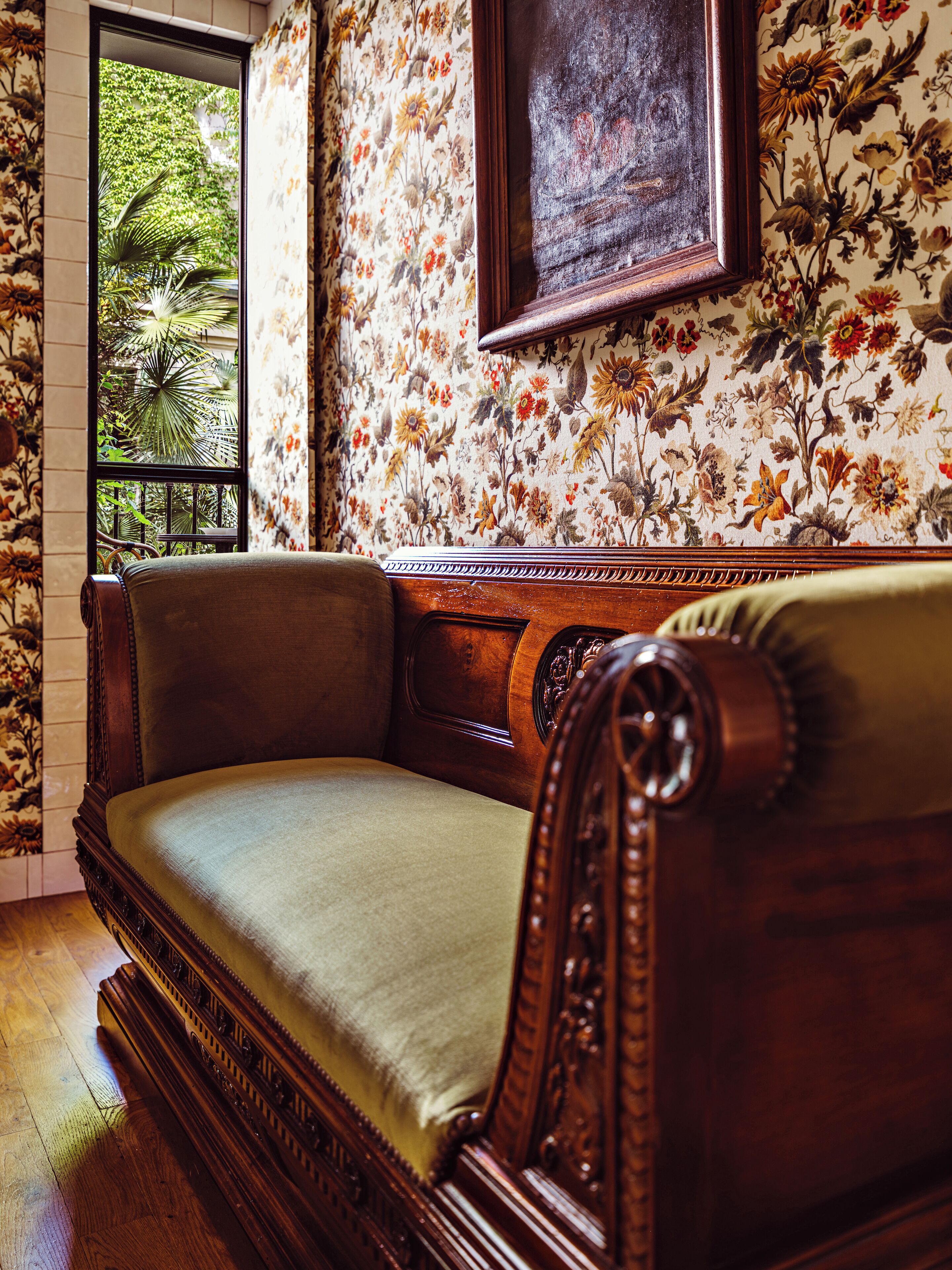 A vintage-style sofa with a cushioned seat and wooden frame, positioned in a room with floral wallpaper and a large window offering a view of greenery.