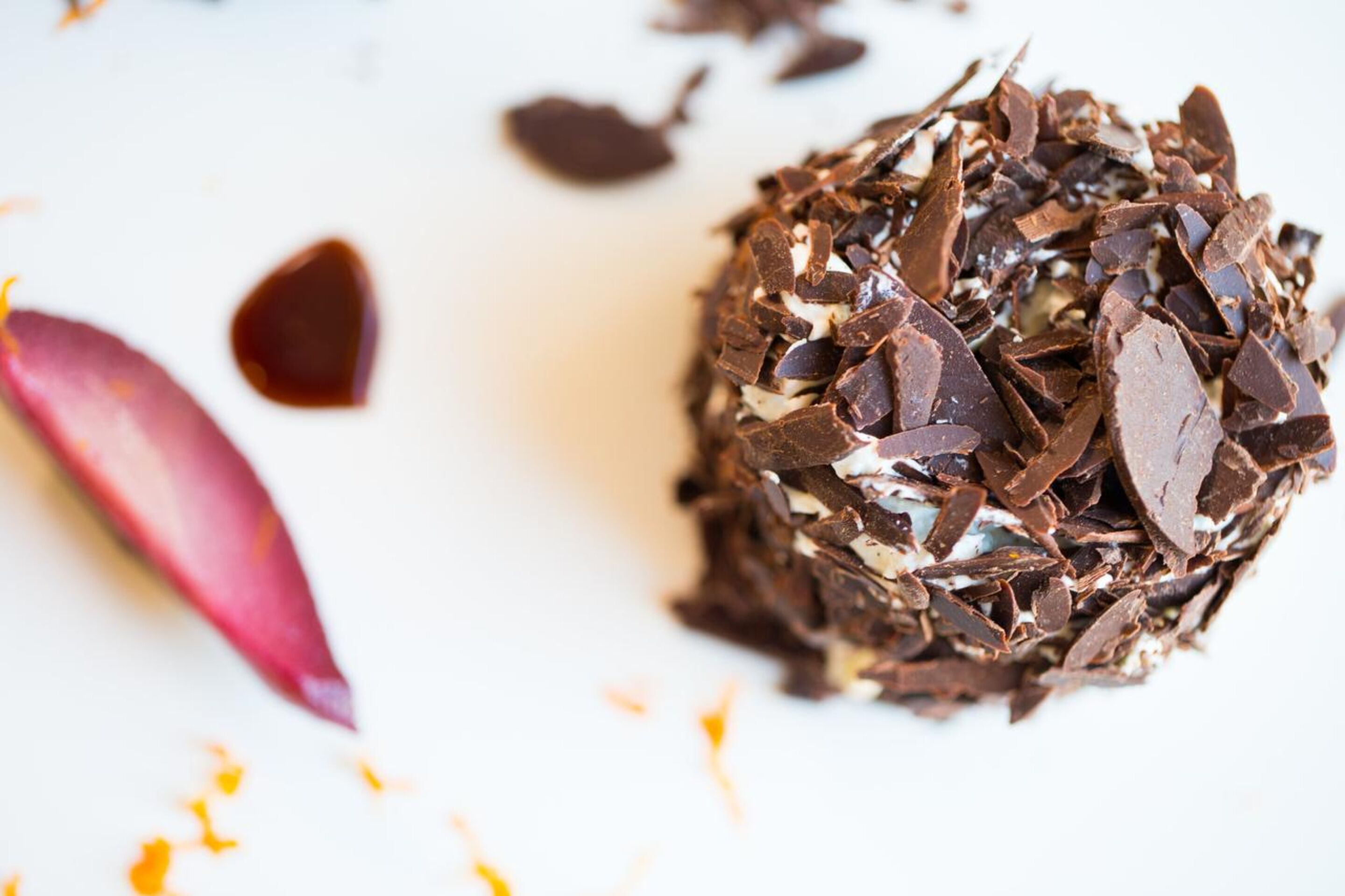 Un postre bañado en chocolate, con virutas de chocolate y una hoja rosa sobre una superficie blanca.