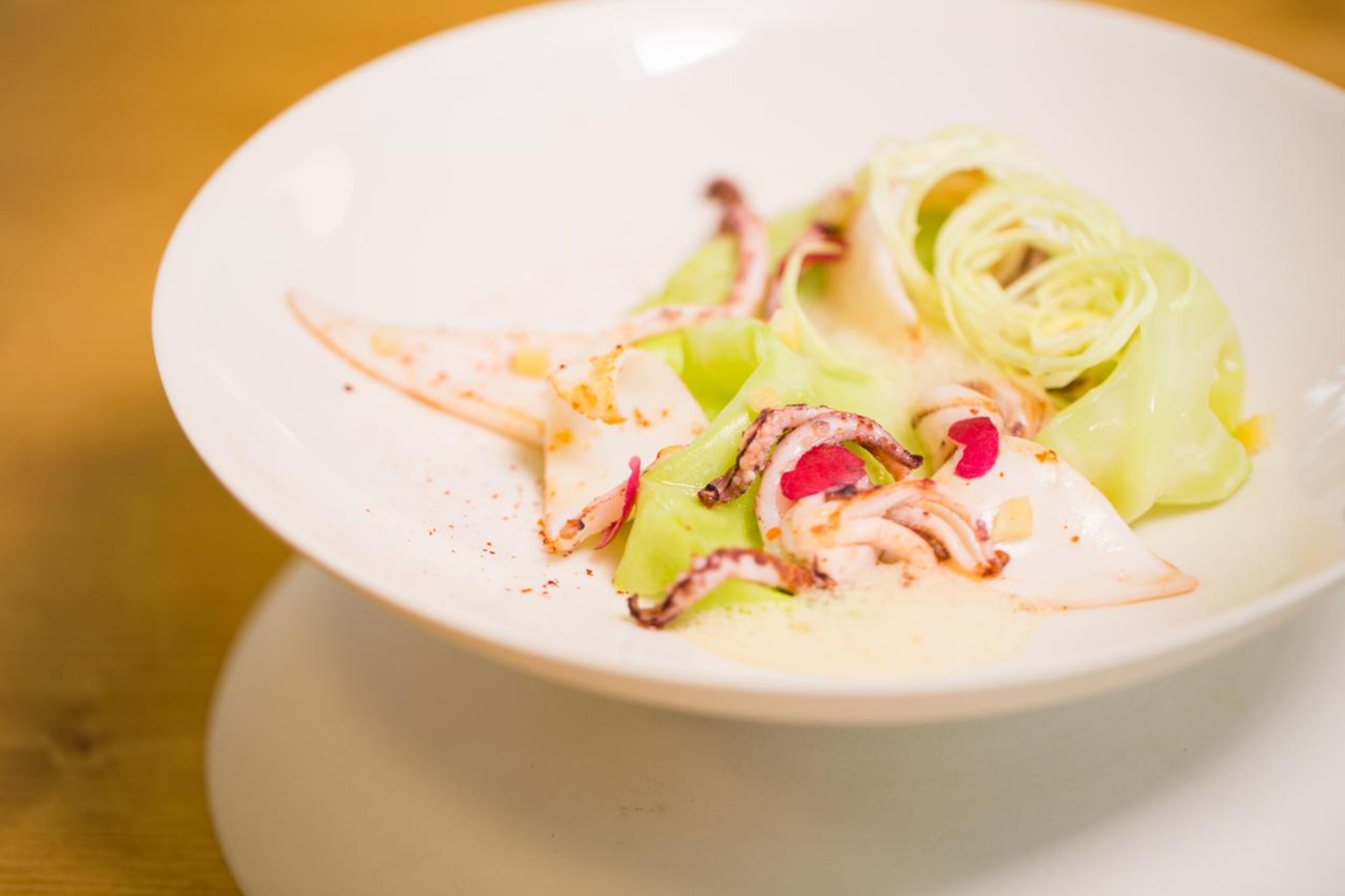 Un plato blanco con un guiso de mariscos, incluyendo calamares y verduras verdes, sobre una mesa de madera.
