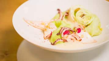 Un plato blanco con un guiso de mariscos, incluyendo calamares y verduras verdes, sobre una mesa de madera.