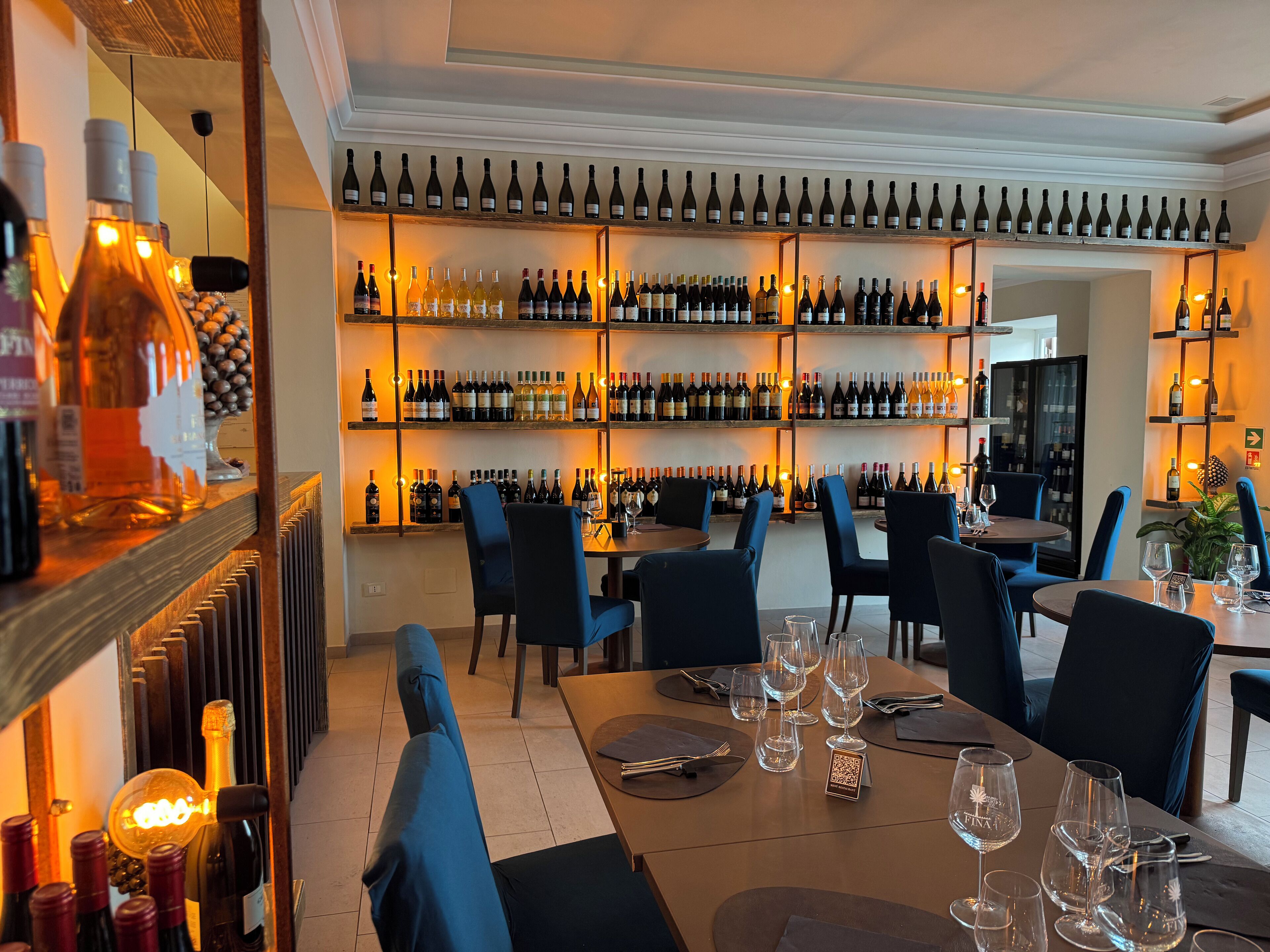 Un interior de restaurante con varias mesas, sillas y una pared llena de botellas de vino.