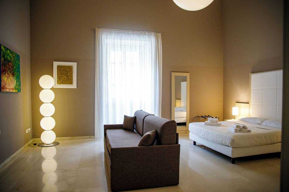 Habitación de hotel con sofá, cama, lámpara de pie y un gran ventanal con cortinas translúcidas.