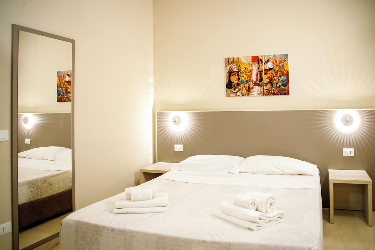 Habitación de hotel con una cama grande, dos mesitas de noche, un espejo e iluminación en la pared.
