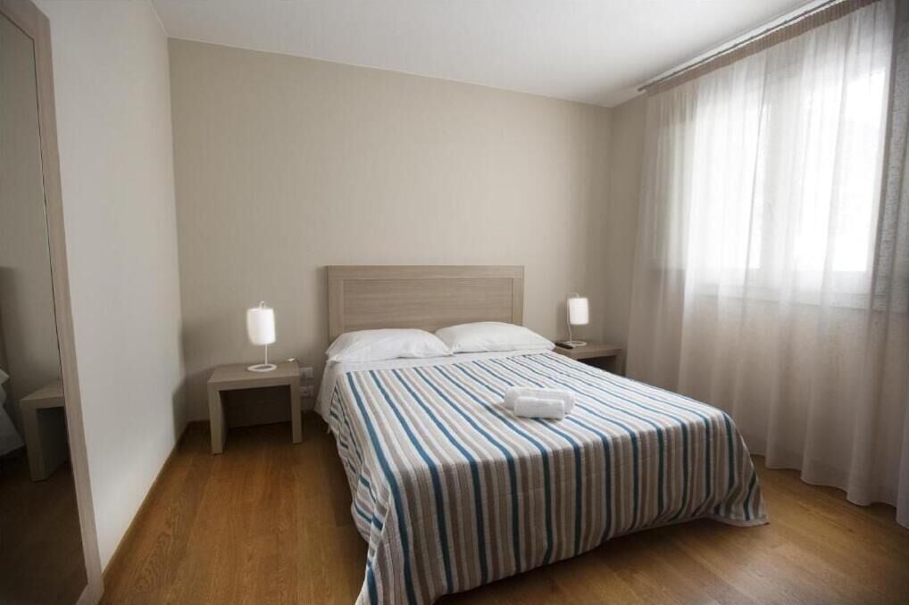 A hotel room with a bed, two bedside tables, a nightstand, a lamp, and a window with sheer curtains.
