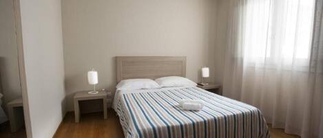 A hotel room with a bed, two bedside tables, a nightstand, a lamp, and a window with sheer curtains.