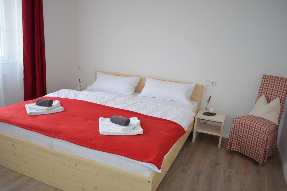 A neatly made bed with a red blanket and two towels, a bedside table with a lamp, and a checkered chair.