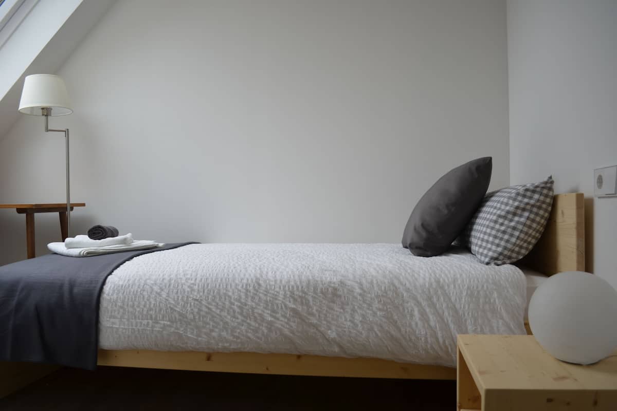 A neatly made bed with a white and gray patterned bedspread, a dark gray pillow, and a checkered pillow on a wooden headboard. A white lamp is on a wooden nightstand beside the bed.