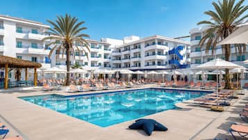 A swimming pool with lounge chairs and umbrellas, surrounded by palm trees and a white building with balconies.