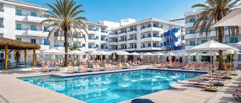 A swimming pool with lounge chairs and umbrellas, surrounded by palm trees and a white building with balconies.