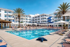 A swimming pool with lounge chairs and umbrellas, surrounded by palm trees and a white building with balconies. - Coral Star Hotel & Apartments (Sant Josep de sa Talaia)