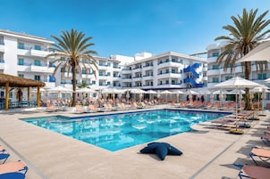 A swimming pool with lounge chairs and umbrellas, surrounded by palm trees and a white building with balconies.