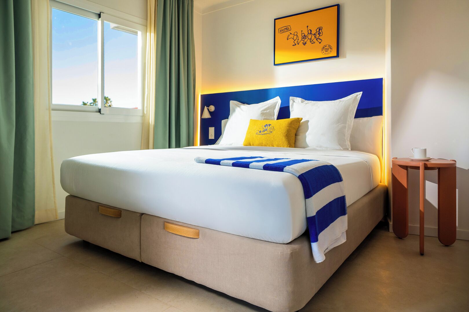 A hotel room with a bed, a bedside table, a window with curtains, and a wall-mounted sign.