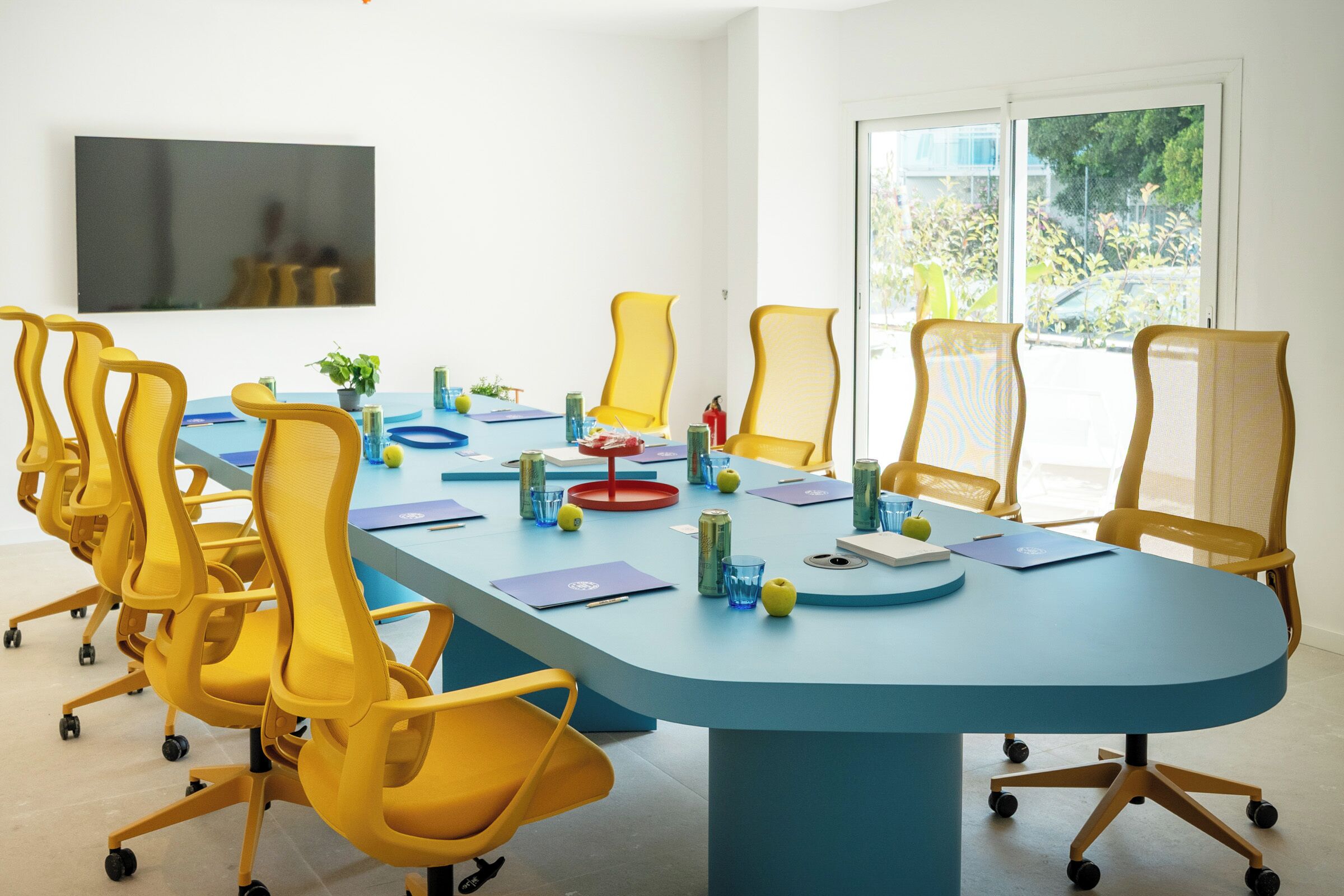 A conference room with a blue table, yellow chairs, and a large window.