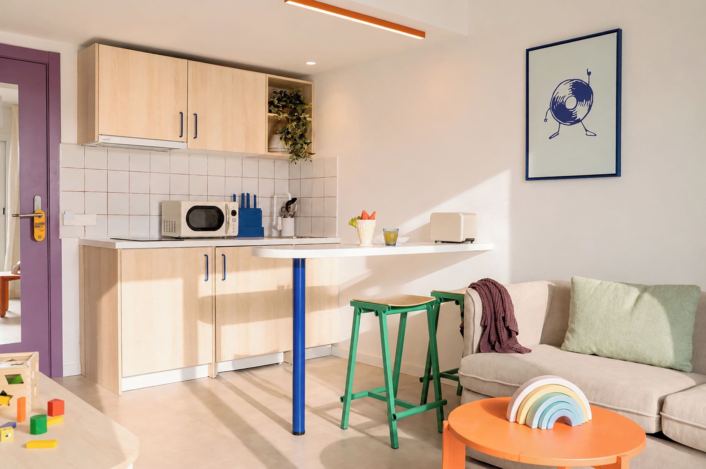 A modern kitchen with wooden cabinets, a microwave, and a white countertop. There is a green stool and a small orange table with a rainbow decoration. A beige sofa and a green cushion are visible in the living area.