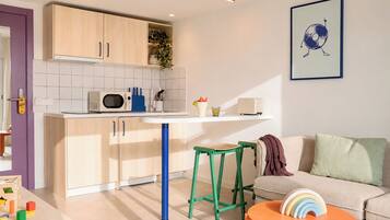 A modern kitchen with wooden cabinets, a microwave, and a white countertop. There is a green stool and a small orange table with a rainbow decoration. A beige sofa and a green cushion are visible in the living area.