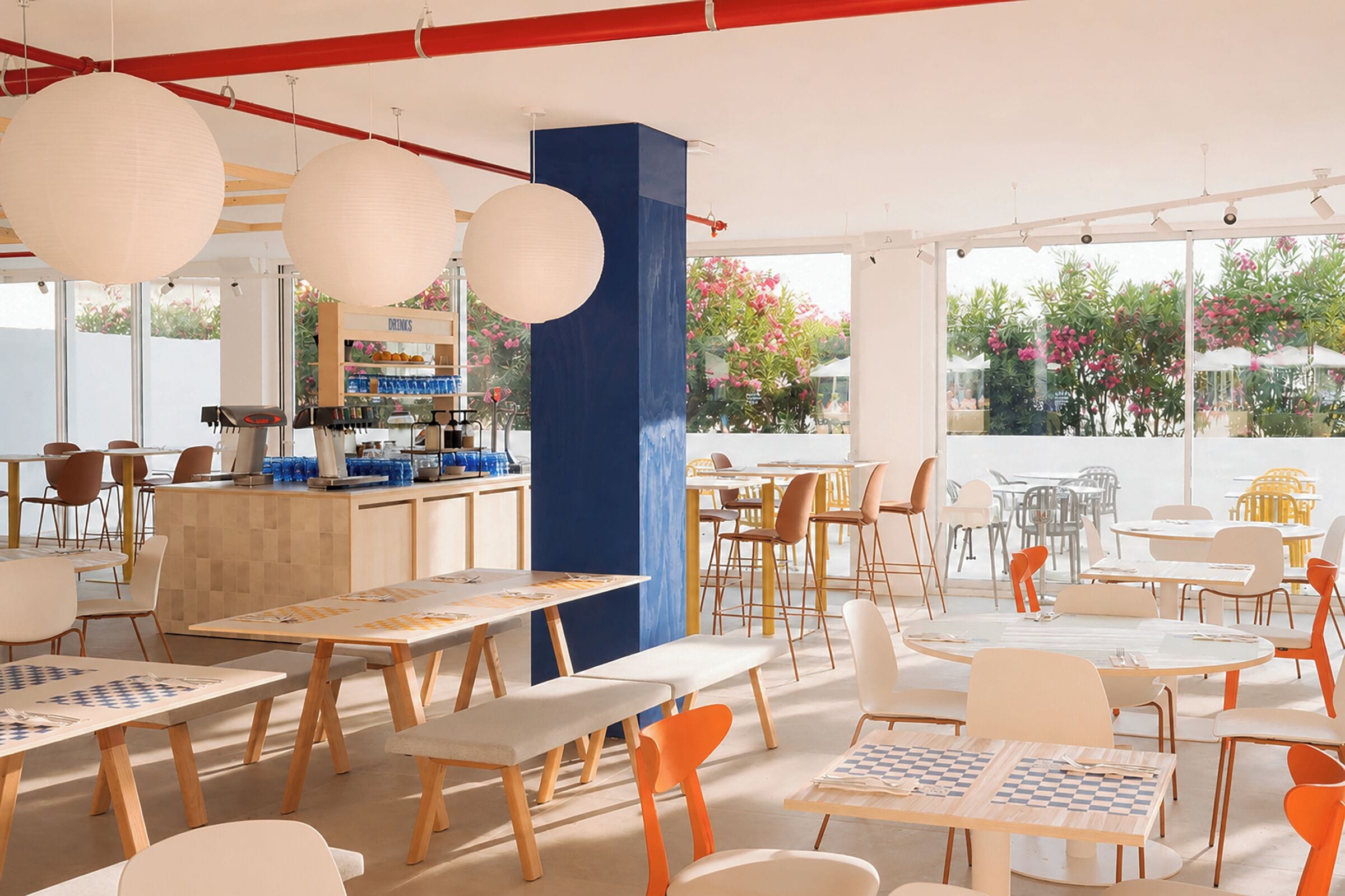 Una cafetería moderna con varias mesas y sillas, una barra y grandes ventanales que ofrecen vistas al exterior.