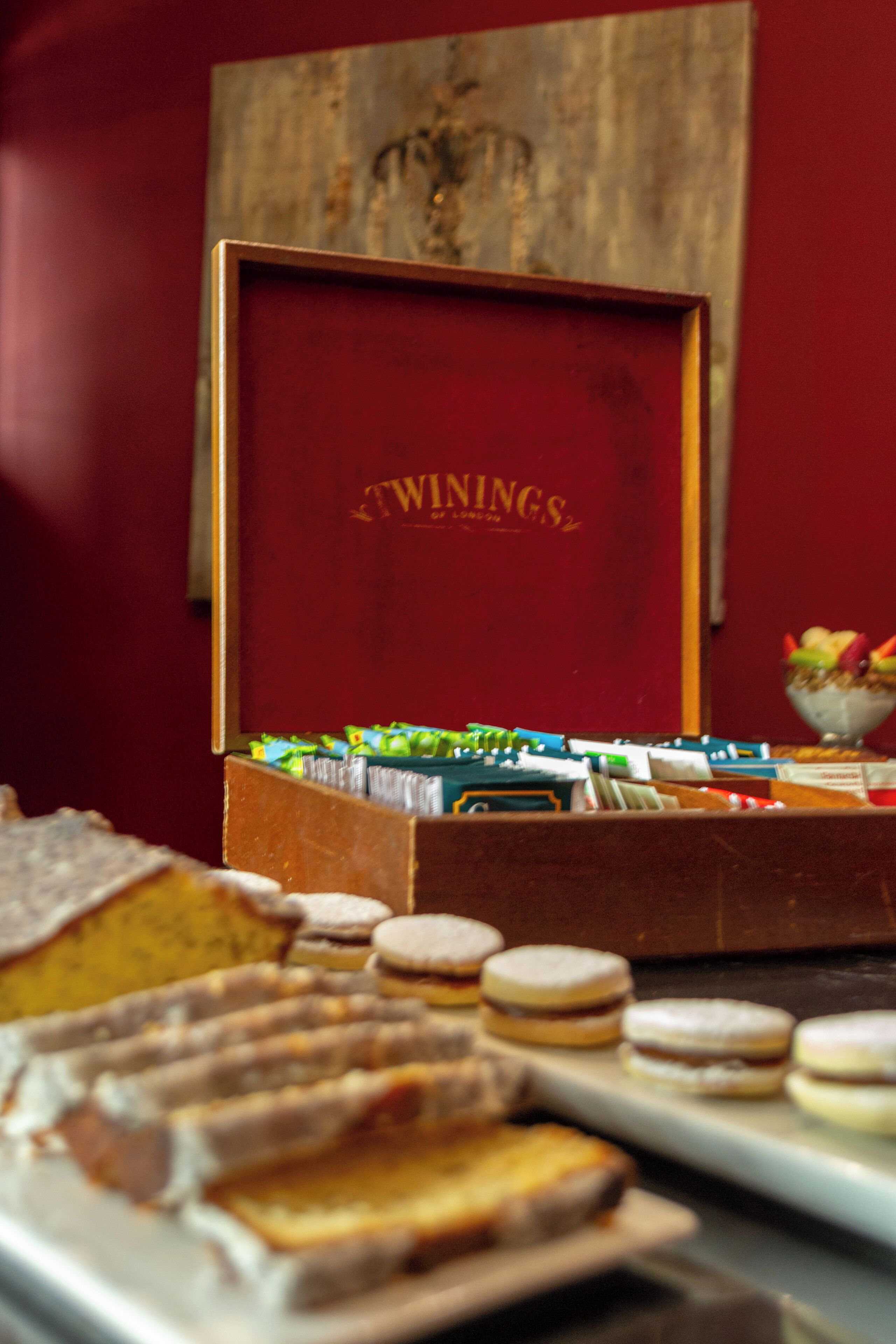 Uma mesa com doces, incluindo diversos pastéis, e uma caixa vermelha com a inscrição "TWININGS" ao fundo.