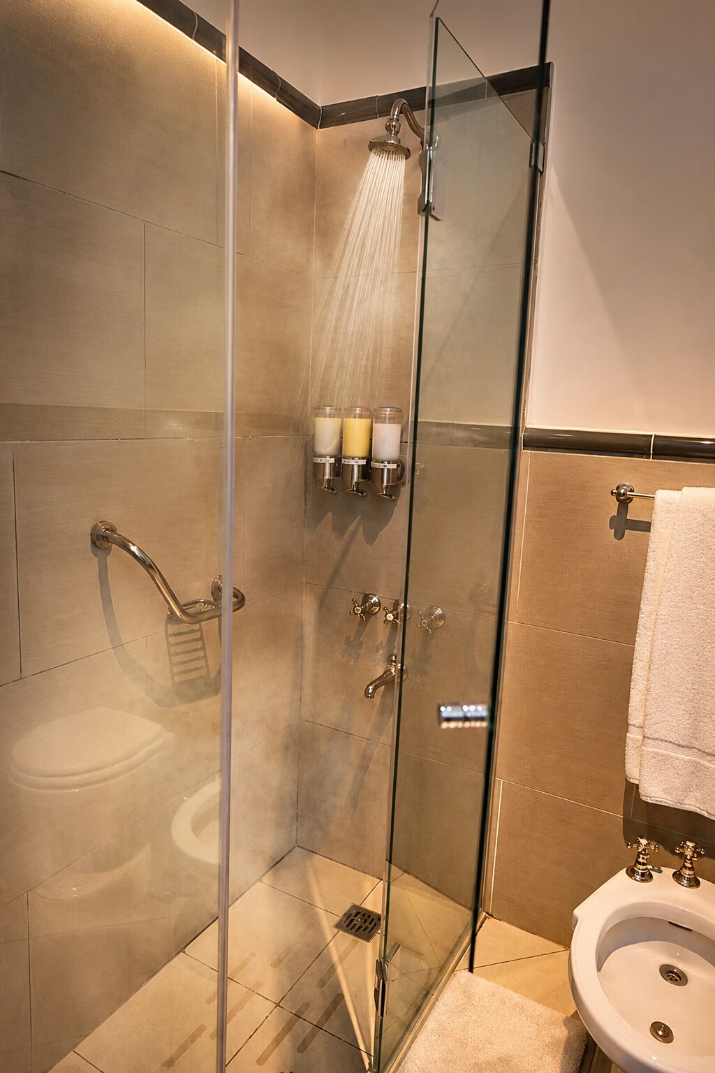 A glass-enclosed shower with a handheld showerhead, a handheld showerhead, and a glass-enclosed sink with a towel hanging on a towel rack.