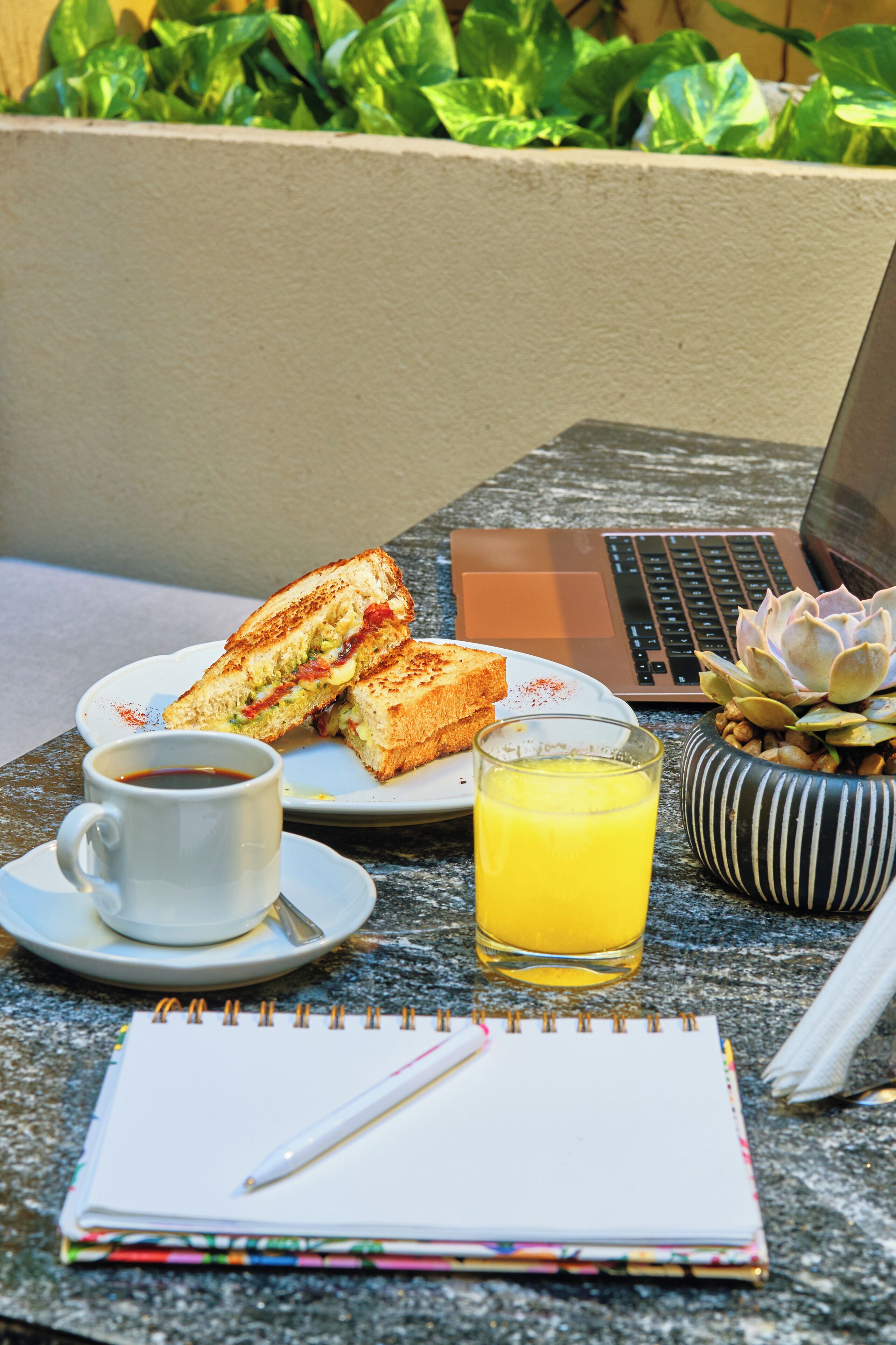 A table with a laptop, a sandwich, a cup of coffee, a glass of orange juice, a notebook, and a pen.