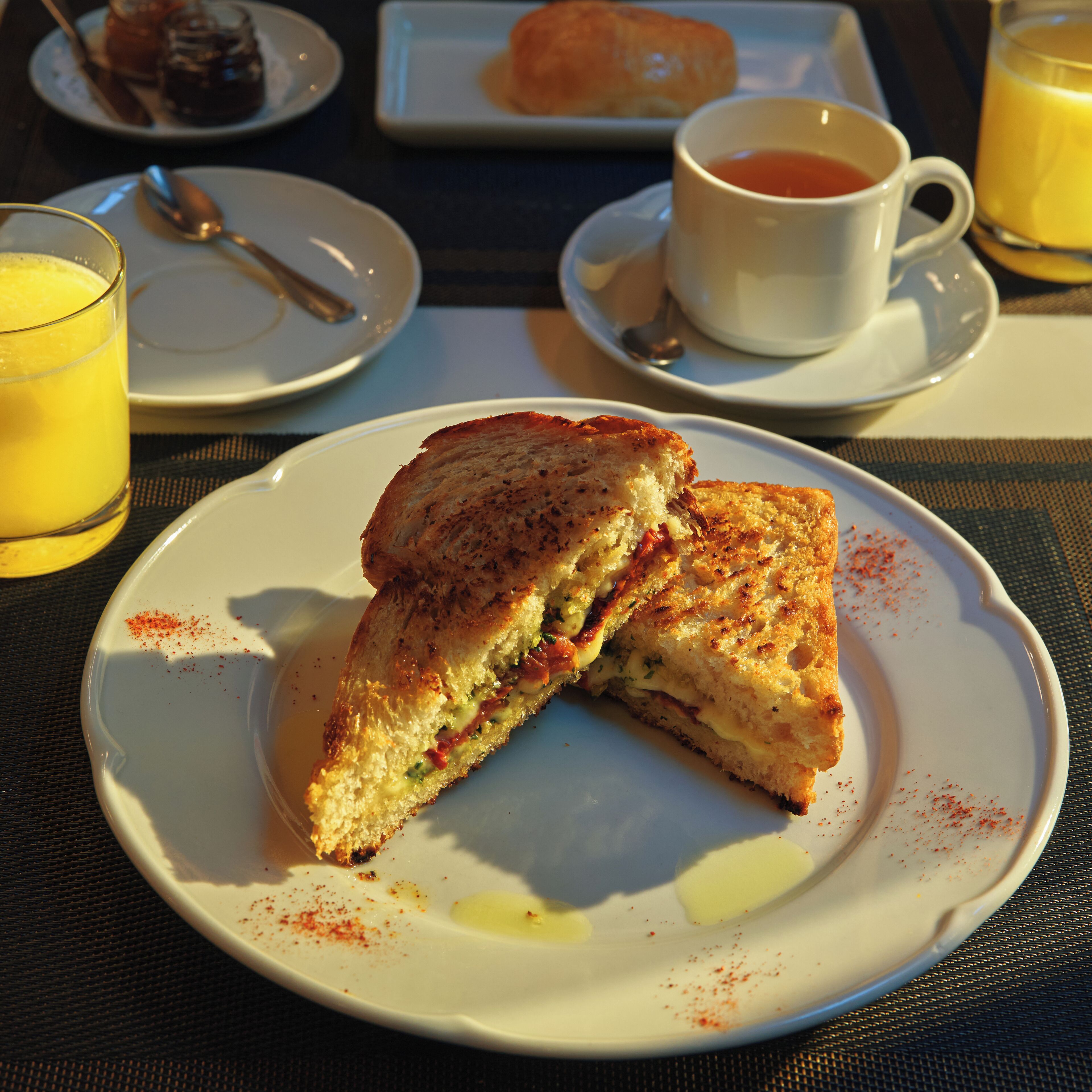 A breakfast setting with a sandwich on a plate, a cup of tea, and a glass of orange juice.