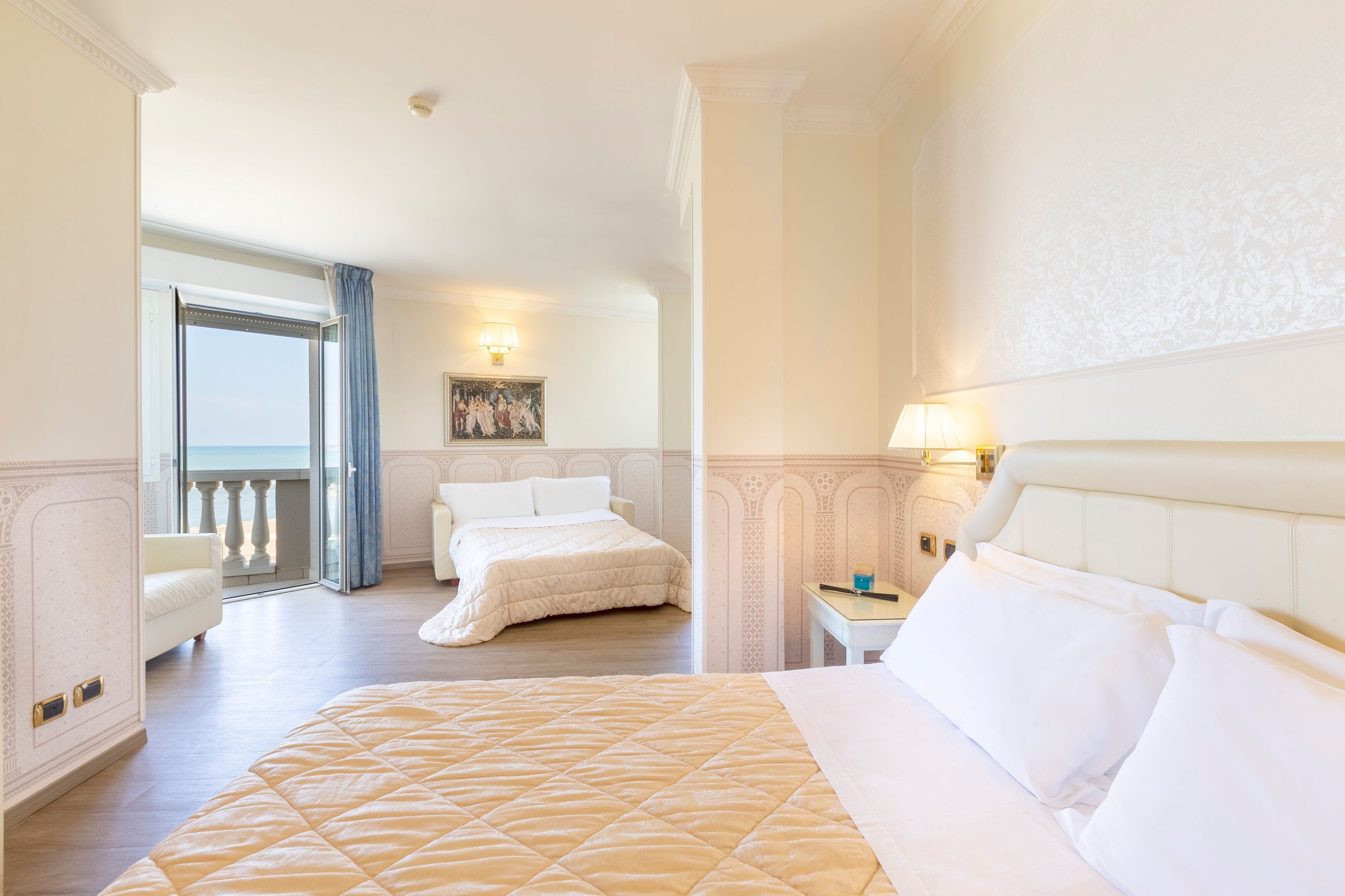 A hotel room with a large bed, a nightstand, a lamp, a painting on the wall, and a view of the sea through an open door.