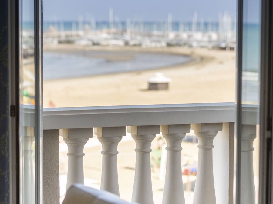 A view from a window with a white balustrade, overlooking a beach with a sandy shore and a pier in the distance.
