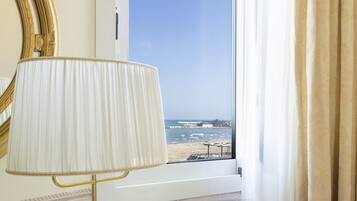 Ein Zimmer mit einem Bett, einem Nachttisch mit Lampe, einem Fenster mit Blick auf den Strand und einem Spiegel an der Wand.