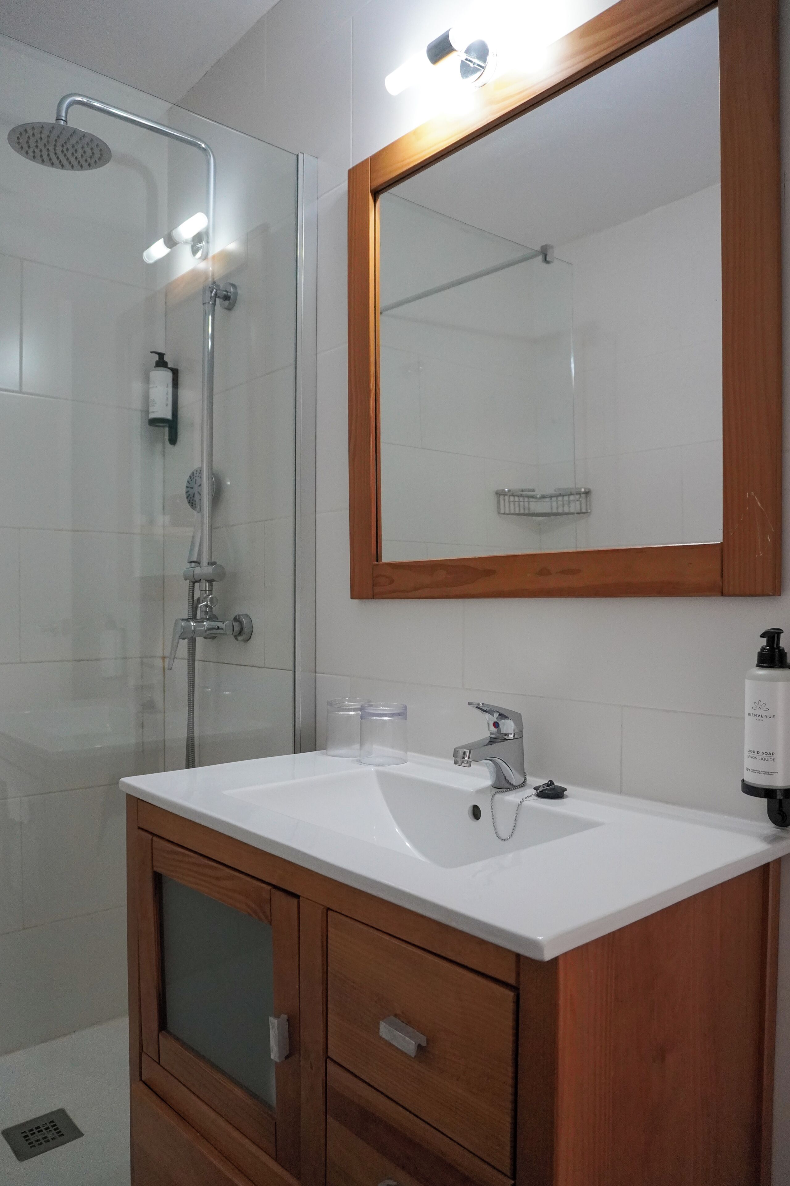 Un baño moderno con ducha de vidrio, tocador de madera con lavamanos y espejo con marco de madera.