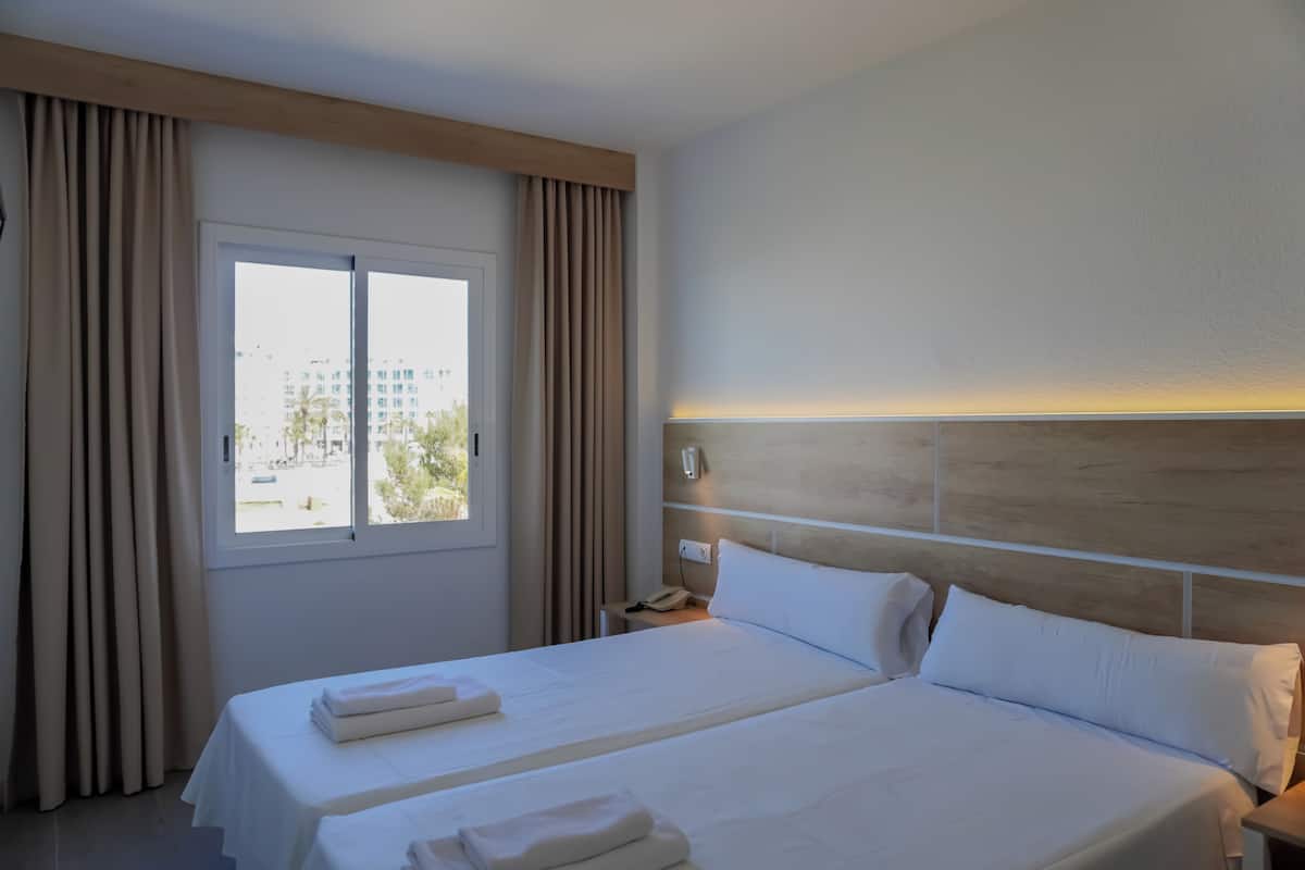 A hotel room with two beds, a window with curtains, a bedside table with a phone, and a view of buildings outside.