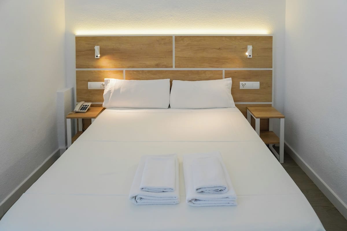 A neatly made bed with white linens and two folded towels, wooden headboard, bedside tables with a phone, and wall-mounted light fixtures.