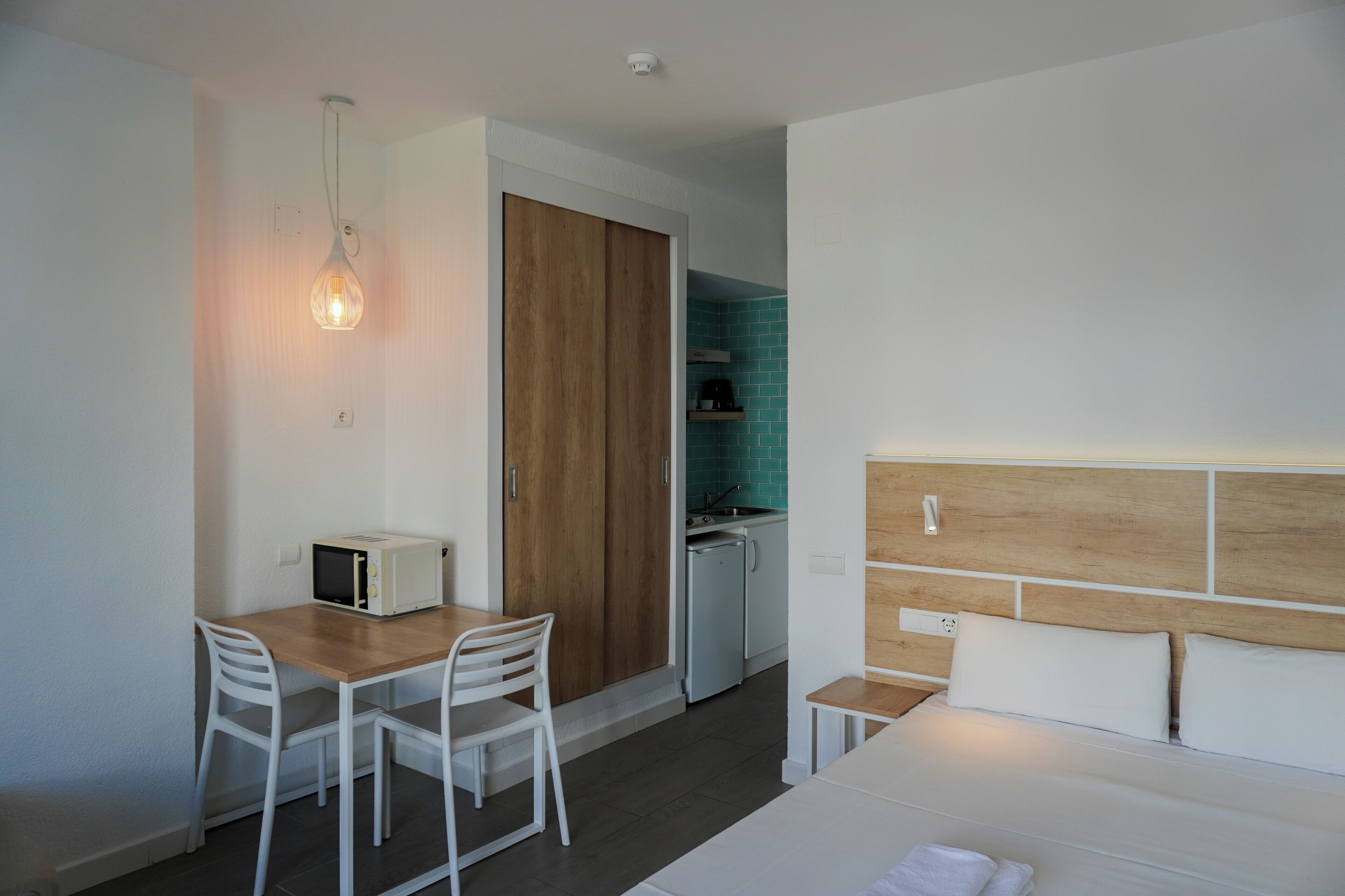 A hotel room with a bed, a small table with chairs, a microwave, a wooden wardrobe, and a kitchenette.