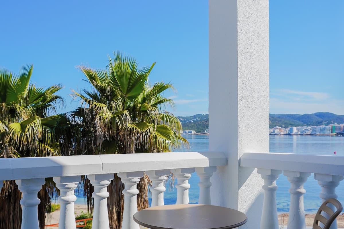A balcony with white balustrades, two chairs, a small round table, and a view of palm trees and a distant cityscape.