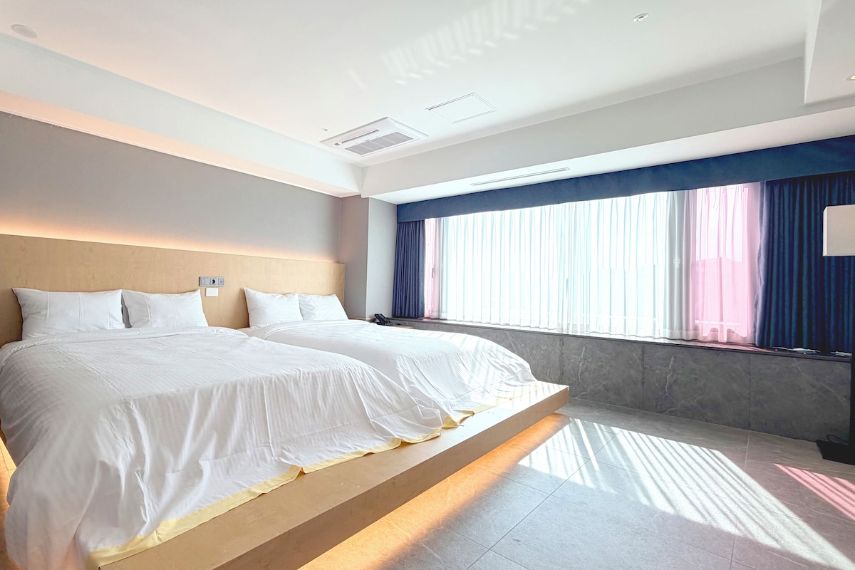 A hotel room with a large bed, a headboard, a bedside table, a telephone, and a window with curtains.