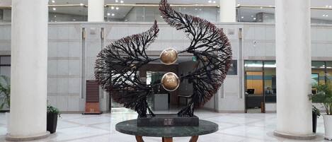 Un vestíbulo con una instalación artística central que presenta una estructura similar a un árbol, con elementos metálicos y una mesa de mármol.