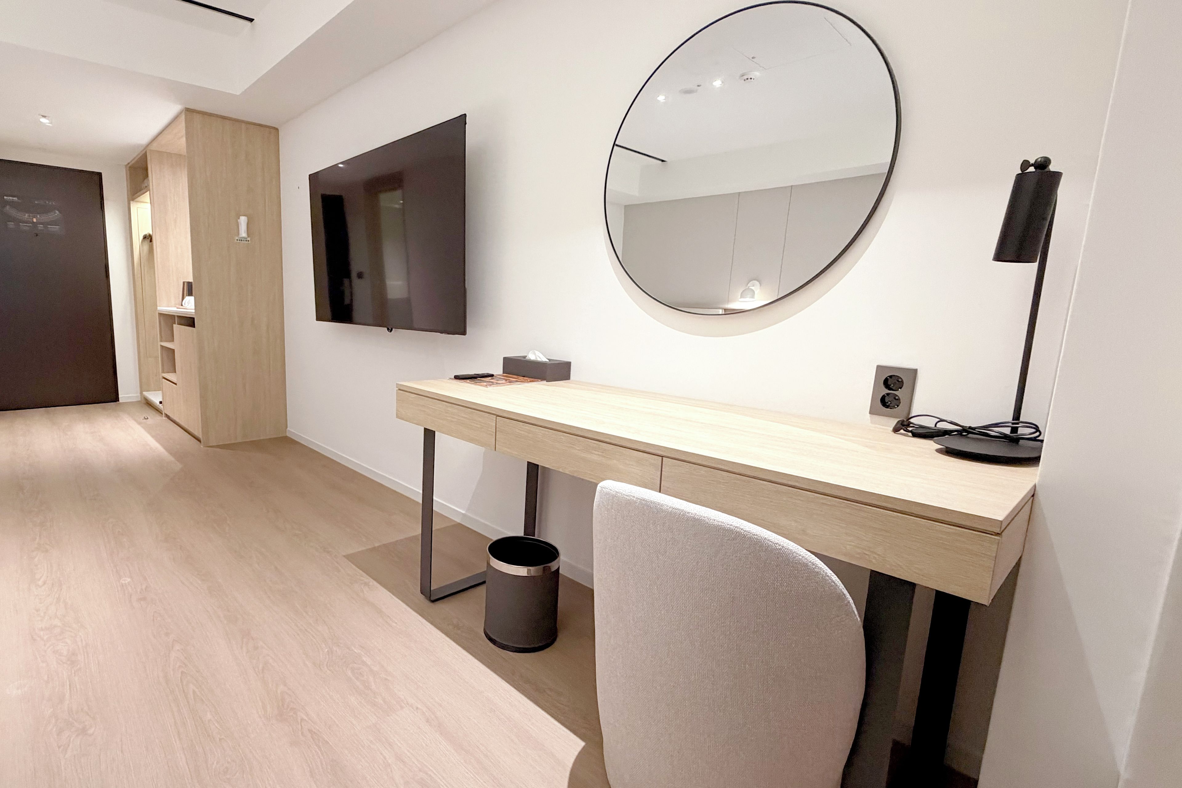A modern bedroom with a wooden desk, a round mirror, a television, and a trash can.