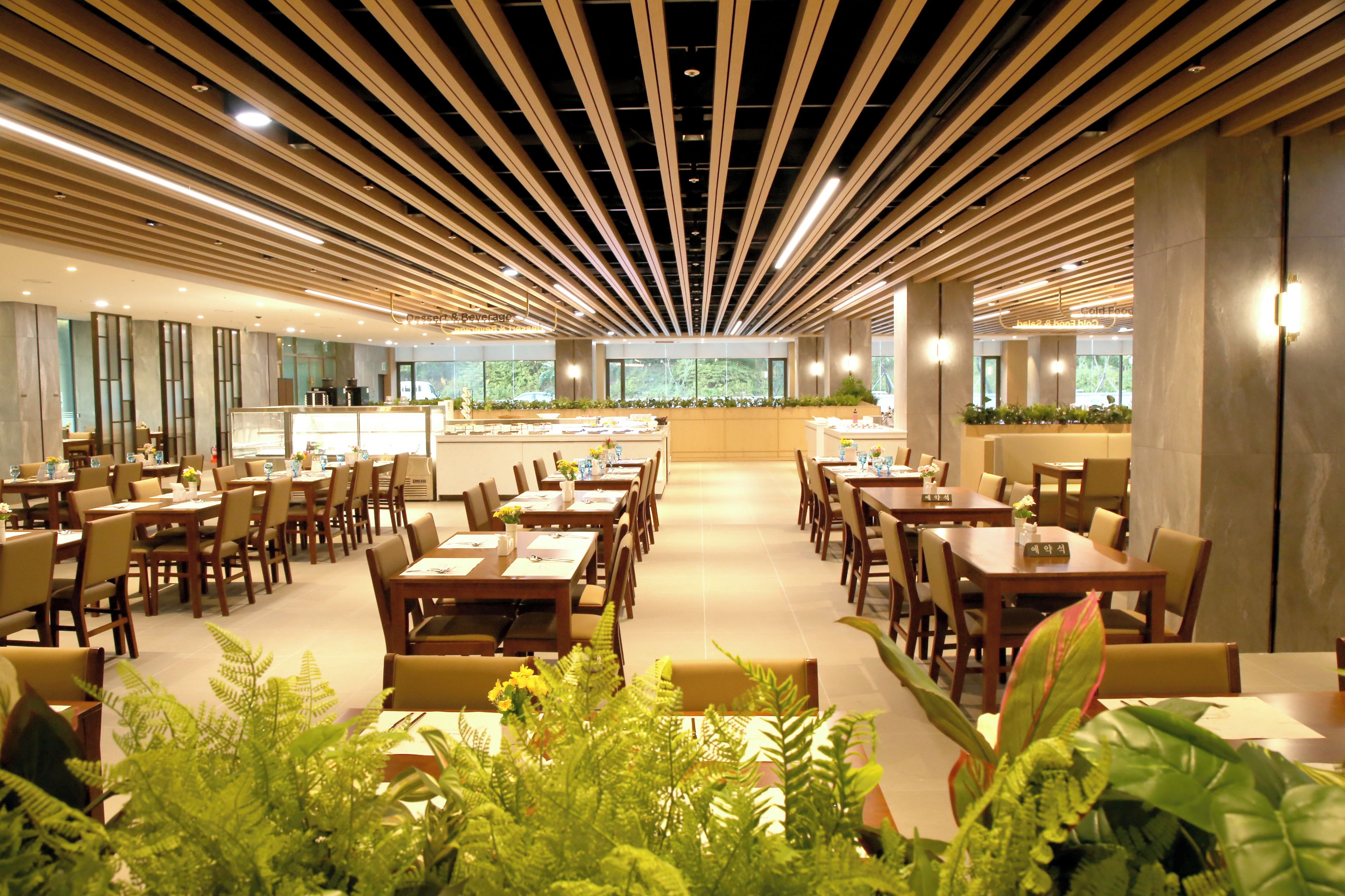 A modern dining area with long tables and chairs, a central counter, and a view of greenery outside.